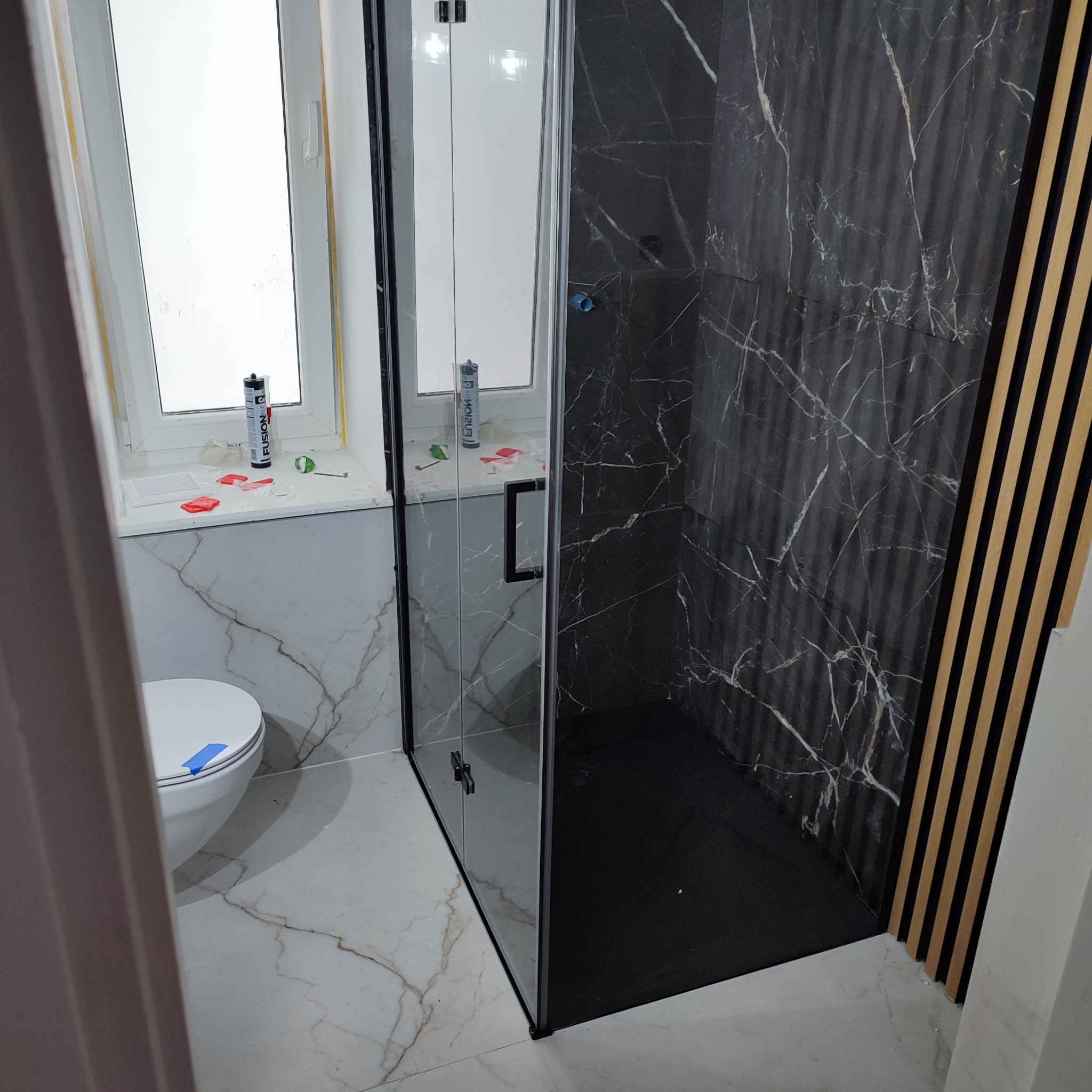 Nowoczesna łazienka po remoncie: kabina prysznicowa z czarnym marmurem, biała toaleta, okno i dekoracyjne lamele na ścianie. Widoczne ślady prac wykończeniowych.