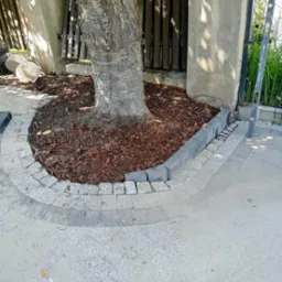 Obrzeże z kostki granitowej wokół pnia drzewa, wysypane korą, z fragmentem ułożonej nawierzchni z kostki brukowej w tle, widoczna poziomica.