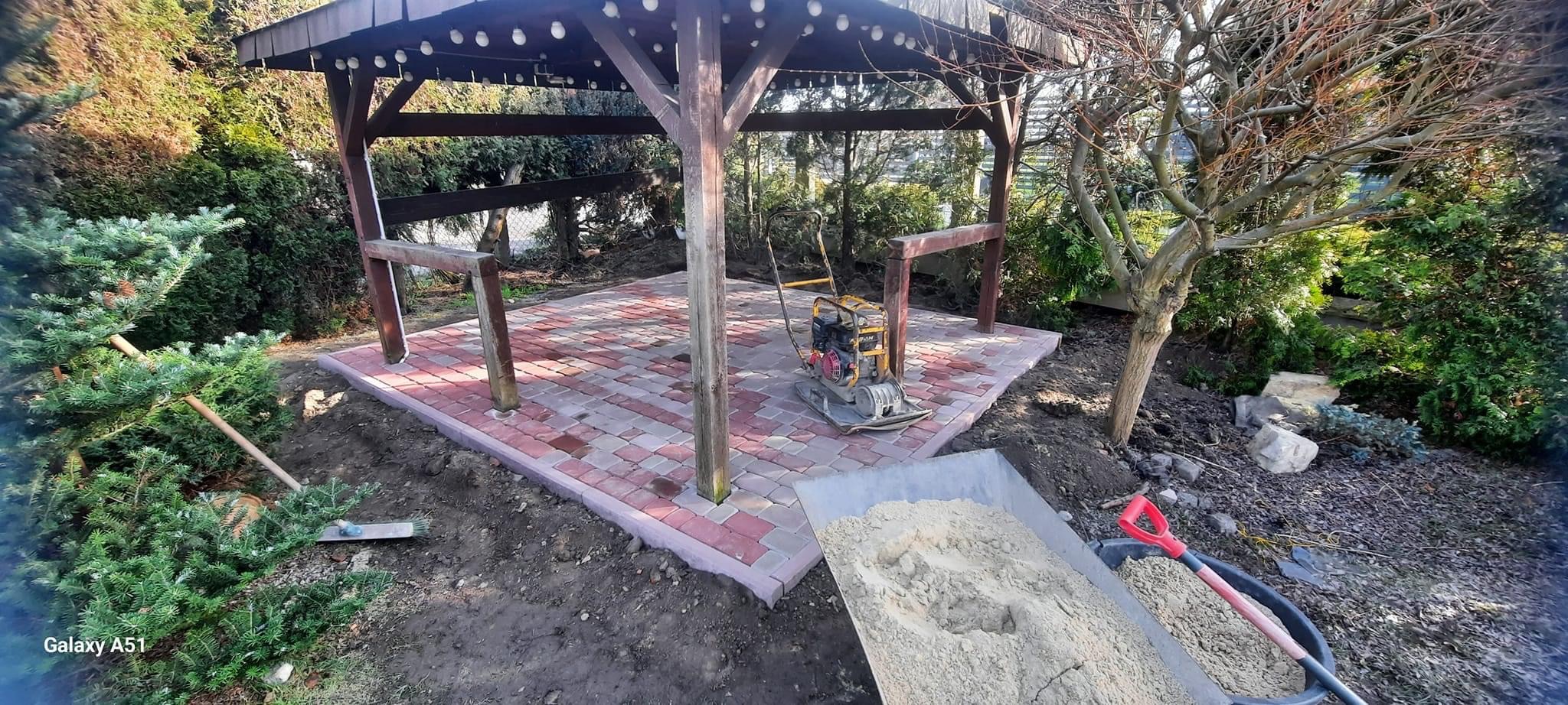 Wykładanie placu pod drewnianą pergolę z czerwono-szarej kostki brukowej, zagęszczarka obok, taczka z piaskiem w tle, w ogrodzie.