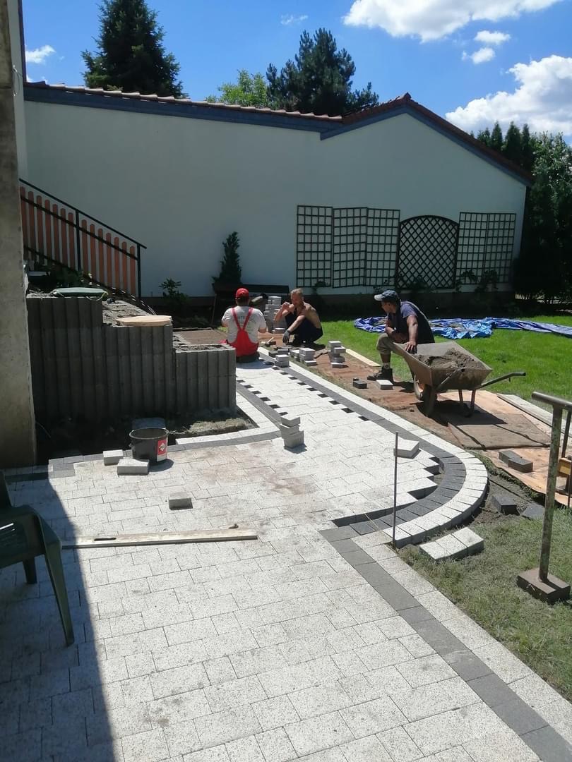 Trzech pracowników układa jasnoszarą kostkę brukową z ciemnoszarymi obrzeżami na posesji z białym budynkiem w tle, widoczna taczka z zaprawą i wiadro.