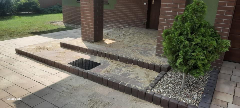 Etap przygotowania podłoża pod taras lub ścieżkę z kostki, widoczny piasek, obrzeża z kostki, kratka odpływowa i małe drzewko iglaste.