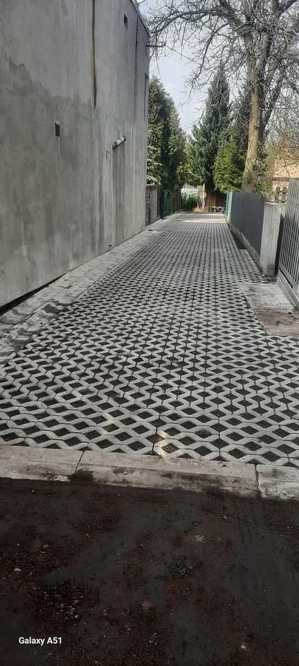 Ułożona szara kostka brukowa ażurowa na podjeździe, widok z perspektywy ulicy, z betonowym obrzeżem, budynkiem po lewej i ogrodzeniem po prawej stronie.