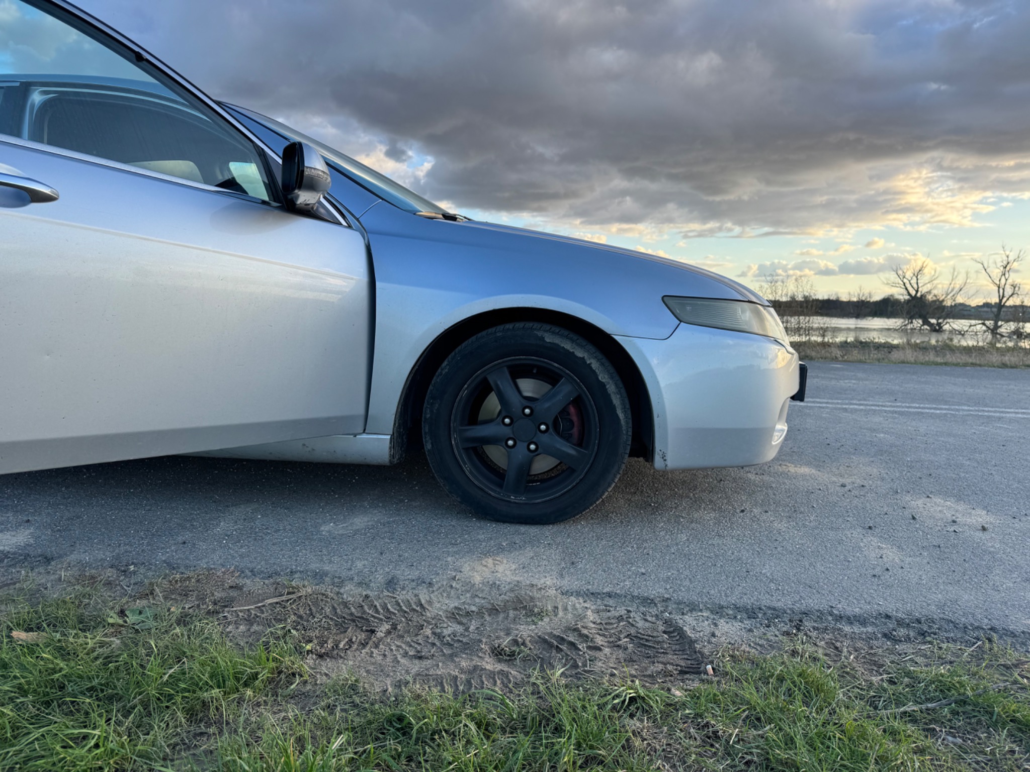 Srebrny samochód z otwartymi drzwiami, czarne felgi, widoczne ślady opon na asfalcie, pochmurne niebo w tle.