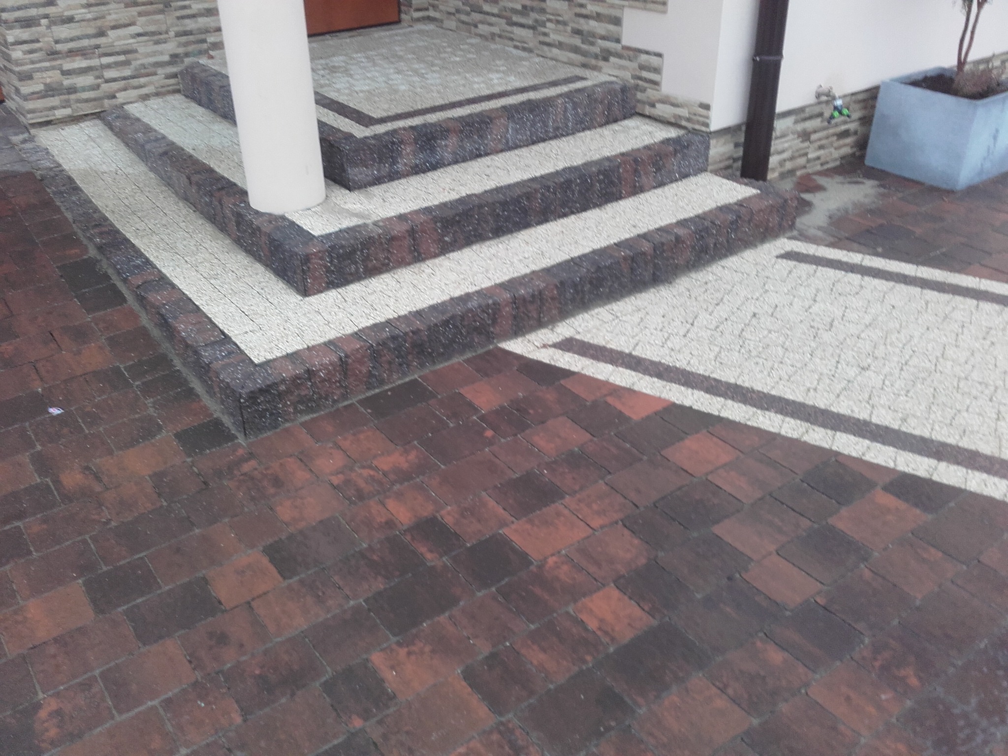Wykończone schody wejściowe z kostki brukowej w dwóch odcieniach, prowadzące do drzwi, z kolumną po lewej stronie i fragmentem brukowanego podjazdu z ciemniejszej kostki.