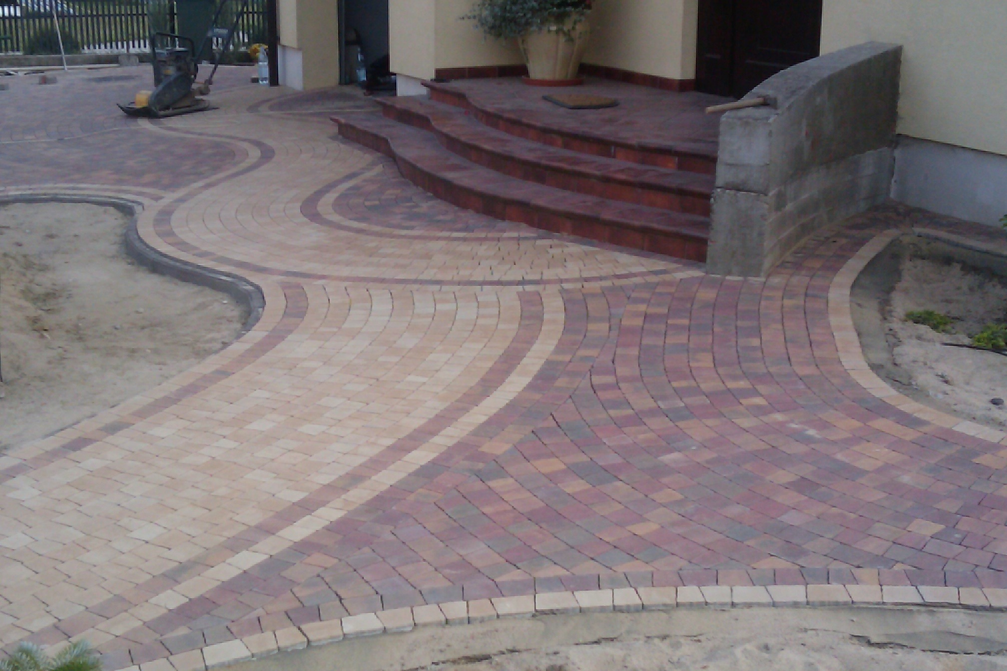 Ukończona ścieżka z kostki brukowej w dwóch odcieniach, prowadząca do schodów wejściowych domu, z widoczną ubijarką w tle i elementami krajobrazu.