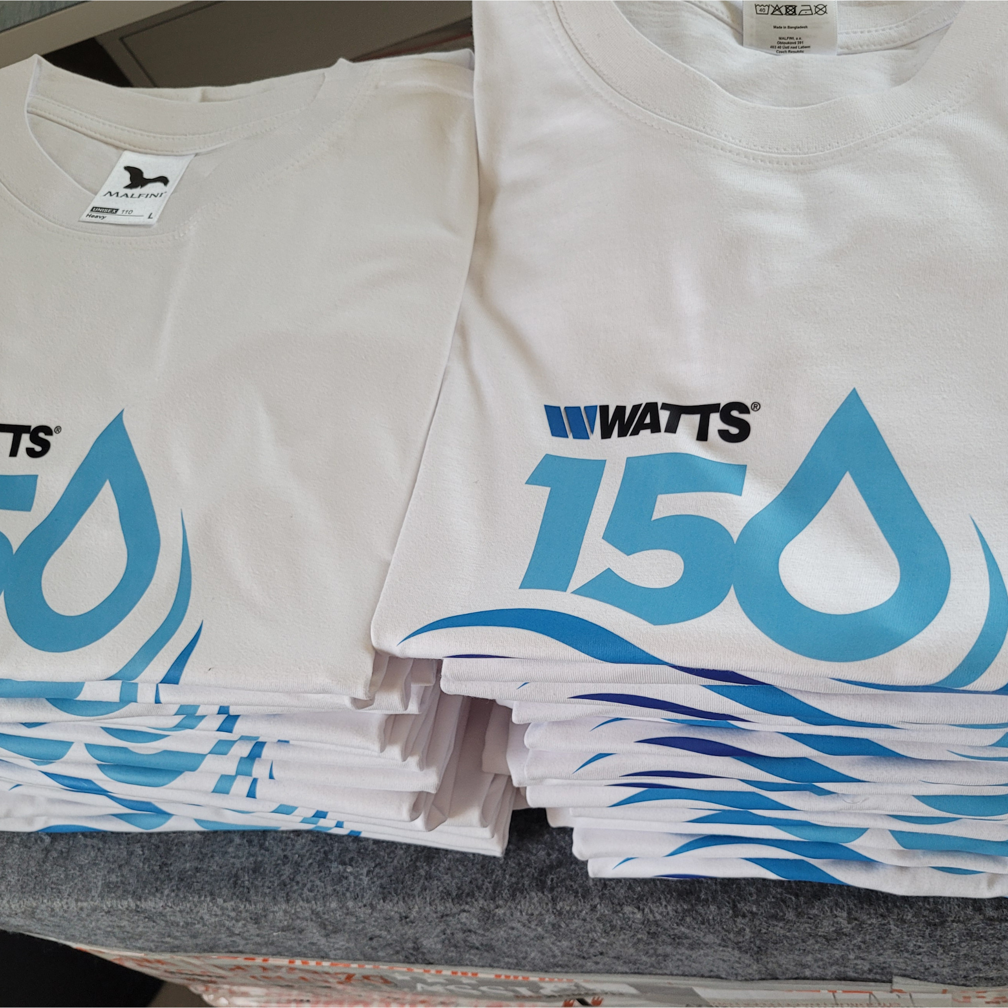 Stos białych koszulek z niebieskim nadrukiem logo 'WATTS 150', widoczne metki producenta Malfini. Prezentacja nadruków DTF Full Kolor na odzieży firmowej.