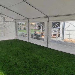 Szubert Events - Wnętrze białego namiotu z widokiem na zewnątrz przez okna z siatką. Podłoga wyłożona zieloną trawą.