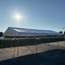 Szubert Events - Rozłożony biały namiot imprezowy na żwirowym podłożu, oświetlony silnym słońcem. W tle zieleń i fragment boiska. Widoczne elementy montażowe.