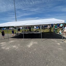 Szubert Events - Biały namiot plenerowy z krzesłami i stołem, ustawiony na asfaltowym placu, w tle widoczni ludzie i fragment boiska. Słoneczny dzień, błękitne niebo.