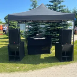 Rozstawiony sprzęt nagłośnieniowy pod czarnym namiotem na trawie, z laptopem na stole. Widoczne głośniki i subwoofery. W tle drzewa i osoby.