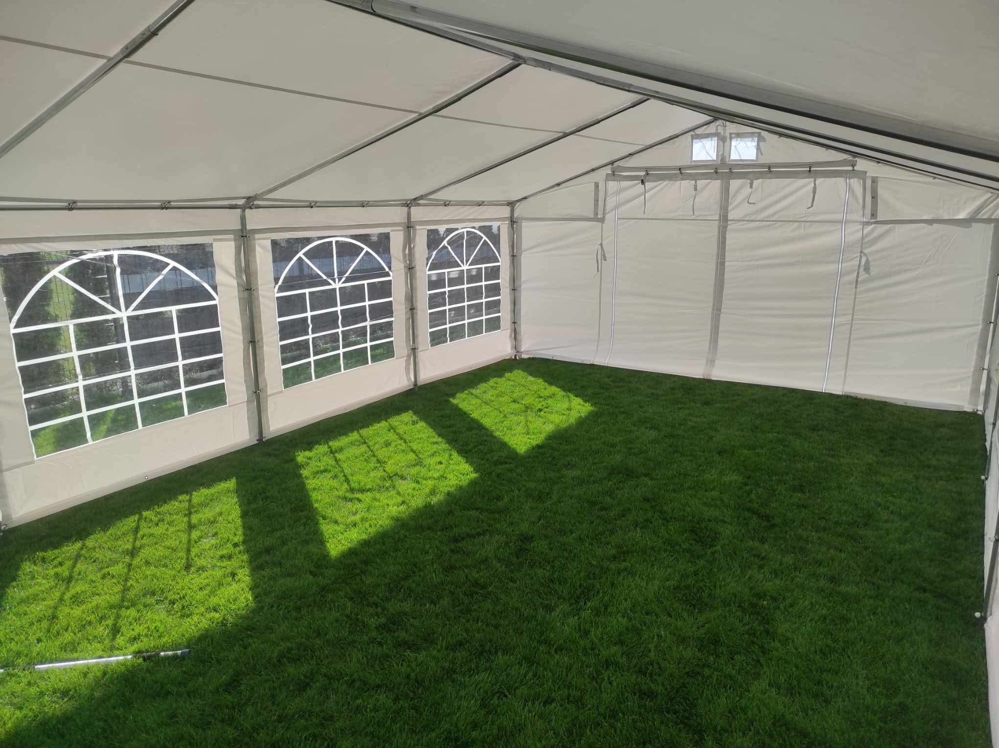 Wnętrze białego namiotu z oknami, zielona trawa wewnątrz, promienie słoneczne tworzą kwadraty na podłożu. Widok od wewnątrz, na podłodze trawa.