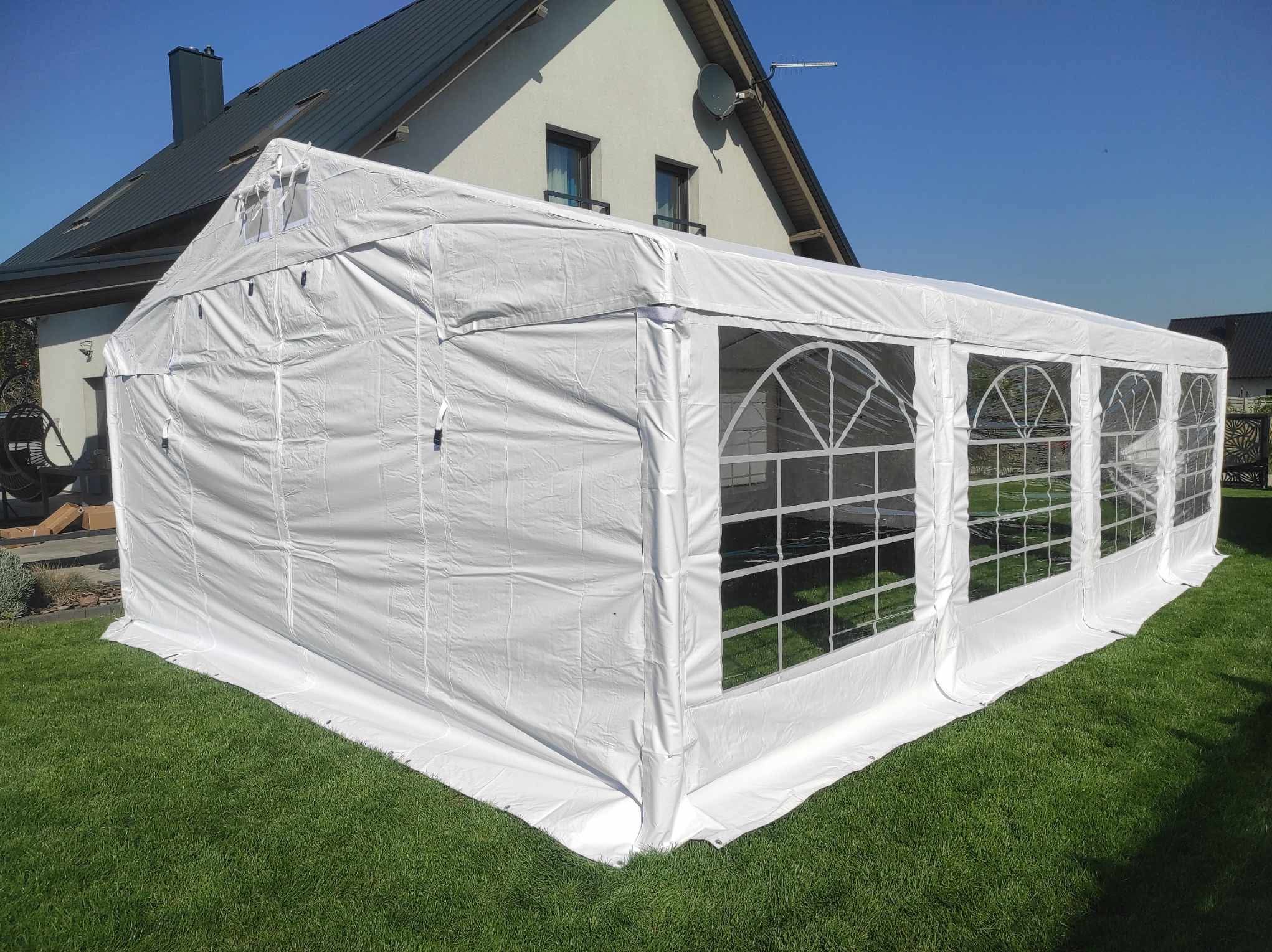 Rozłożony biały namiot imprezowy z oknami na trawniku przed domem jednorodzinnym w słoneczny dzień. Widok z boku na ścianę z oknami.