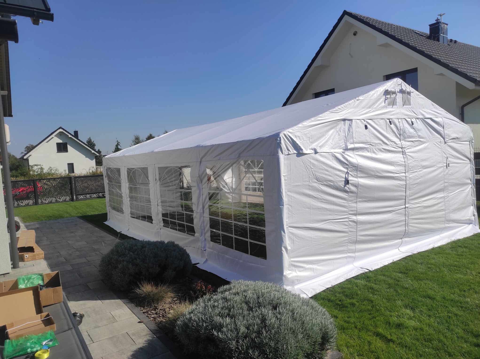Biały namiot imprezowy z oknami ustawiony na trawniku przed domem jednorodzinnym w słoneczny dzień. Widoczne krzewy i fragmenty kostki brukowej.