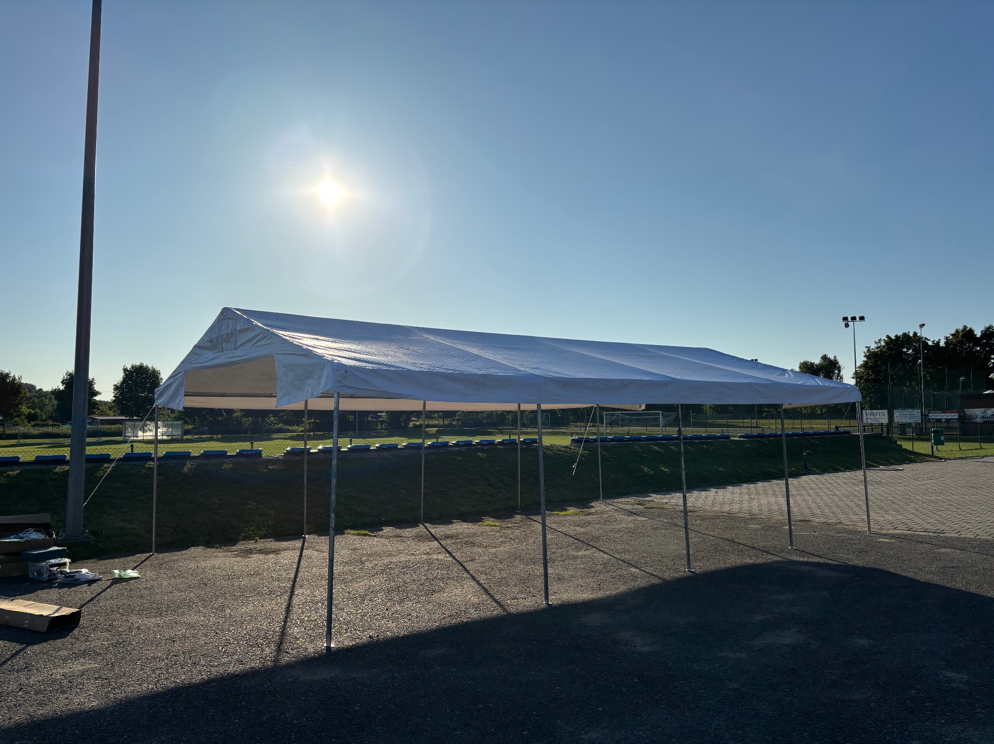 Rozłożony biały namiot imprezowy na żwirowym podłożu, oświetlony silnym słońcem. W tle zieleń i fragment boiska. Widoczne elementy montażowe.