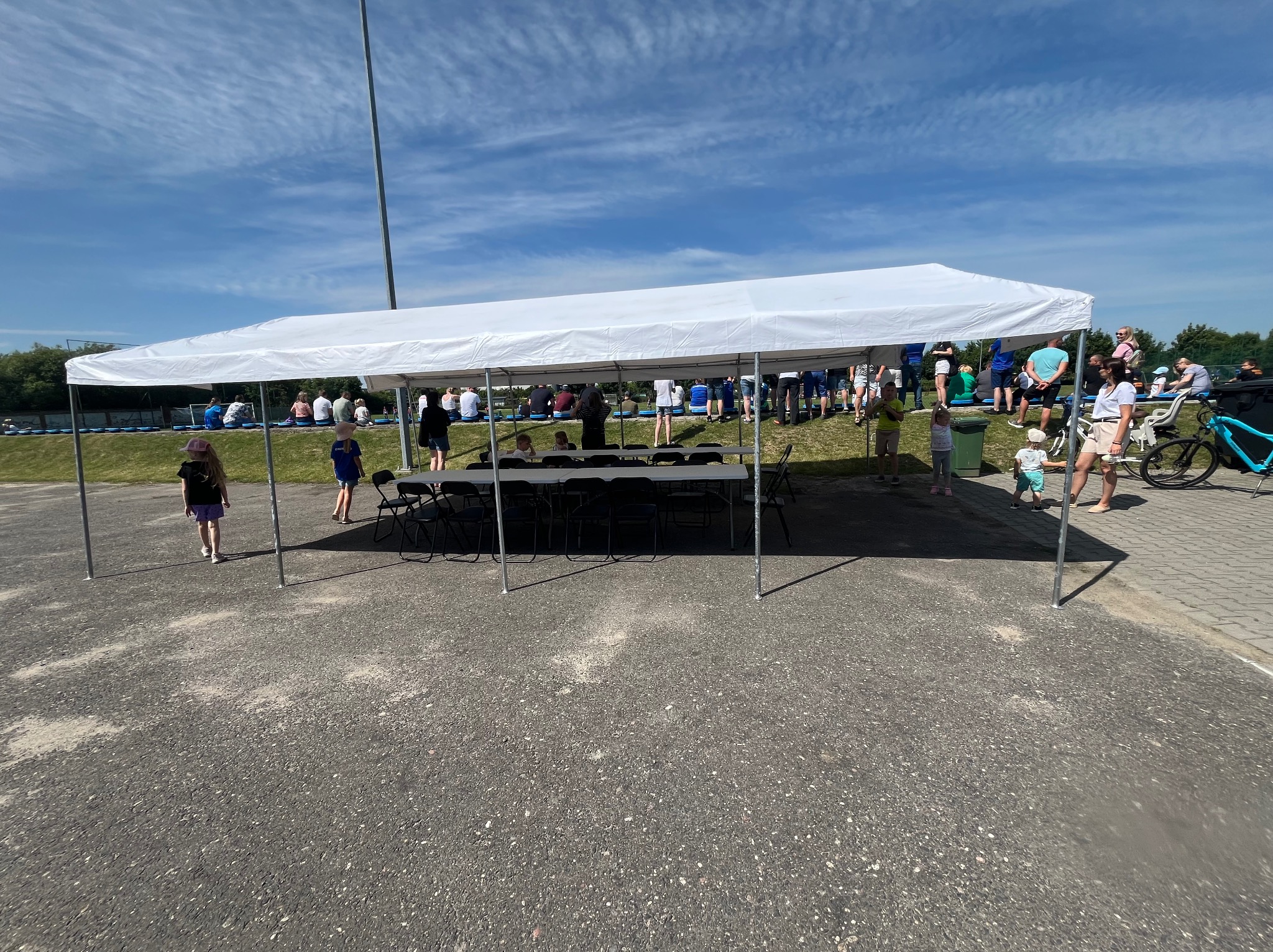 Biały namiot plenerowy z krzesłami i stołem, ustawiony na asfaltowym placu, w tle widoczni ludzie i fragment boiska. Słoneczny dzień, błękitne niebo.