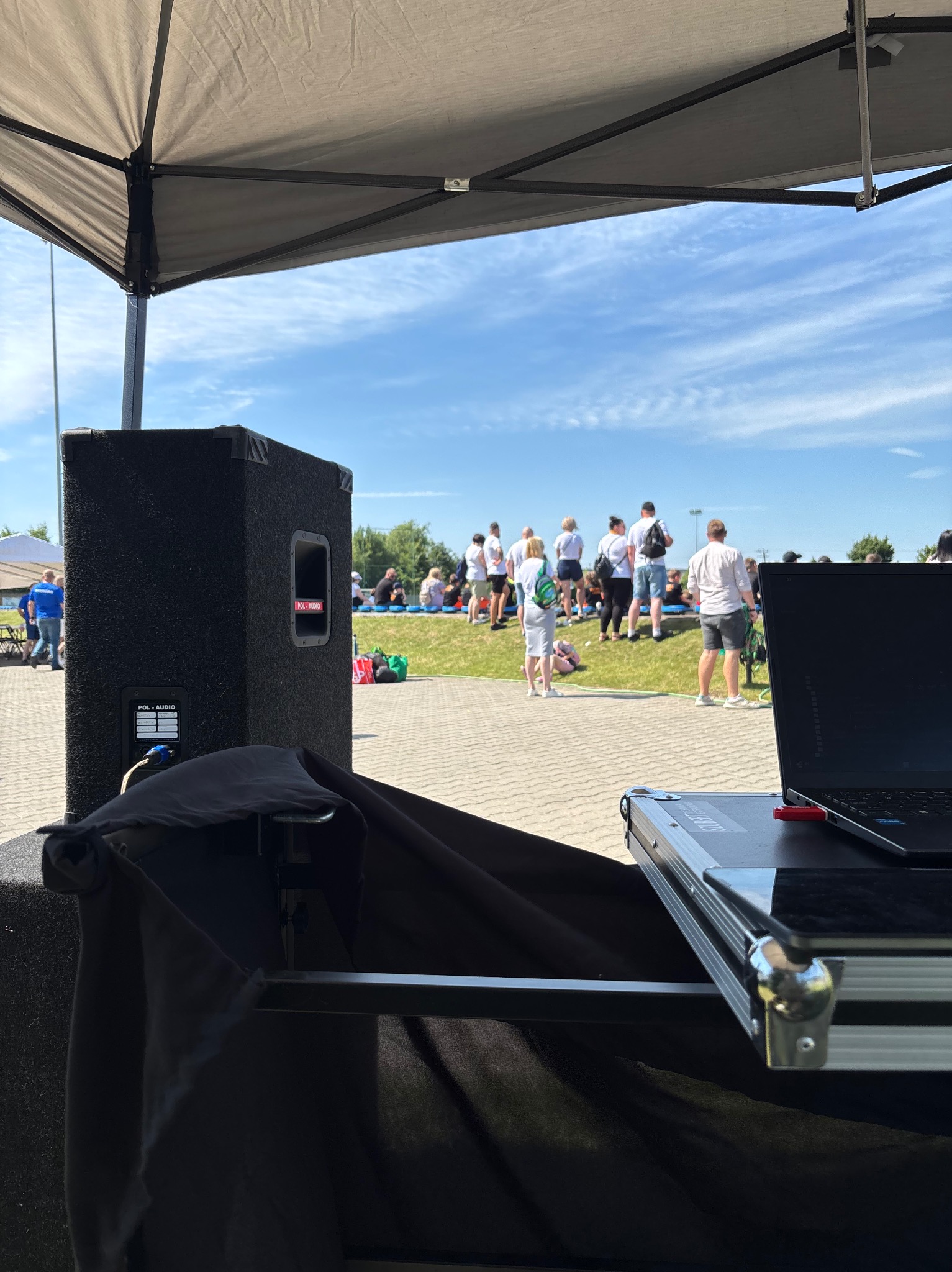 Sprzęt nagłośnieniowy (głośnik, laptop) pod namiotem podczas imprezy plenerowej. W tle grupa osób na trawie.