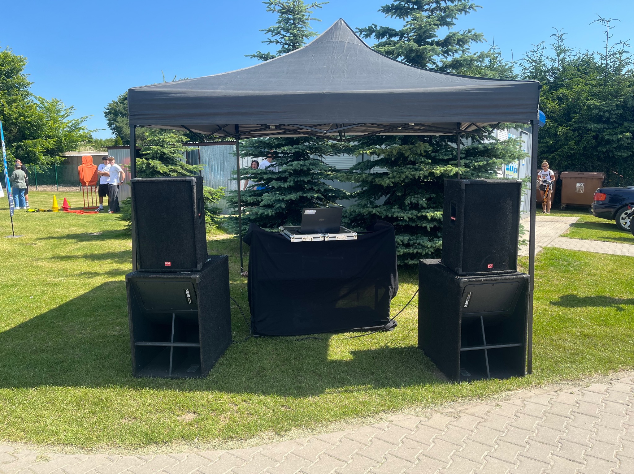 Rozstawiony sprzęt nagłośnieniowy pod czarnym namiotem na trawie, z laptopem na stole. Widoczne głośniki i subwoofery. W tle drzewa i osoby.