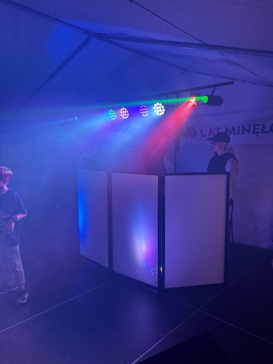 DJ na scenie pod namiotem, oświetlenie sceniczne w kolorach niebieskim, czerwonym i zielonym. Widoczny fragment publiczności po lewej stronie.