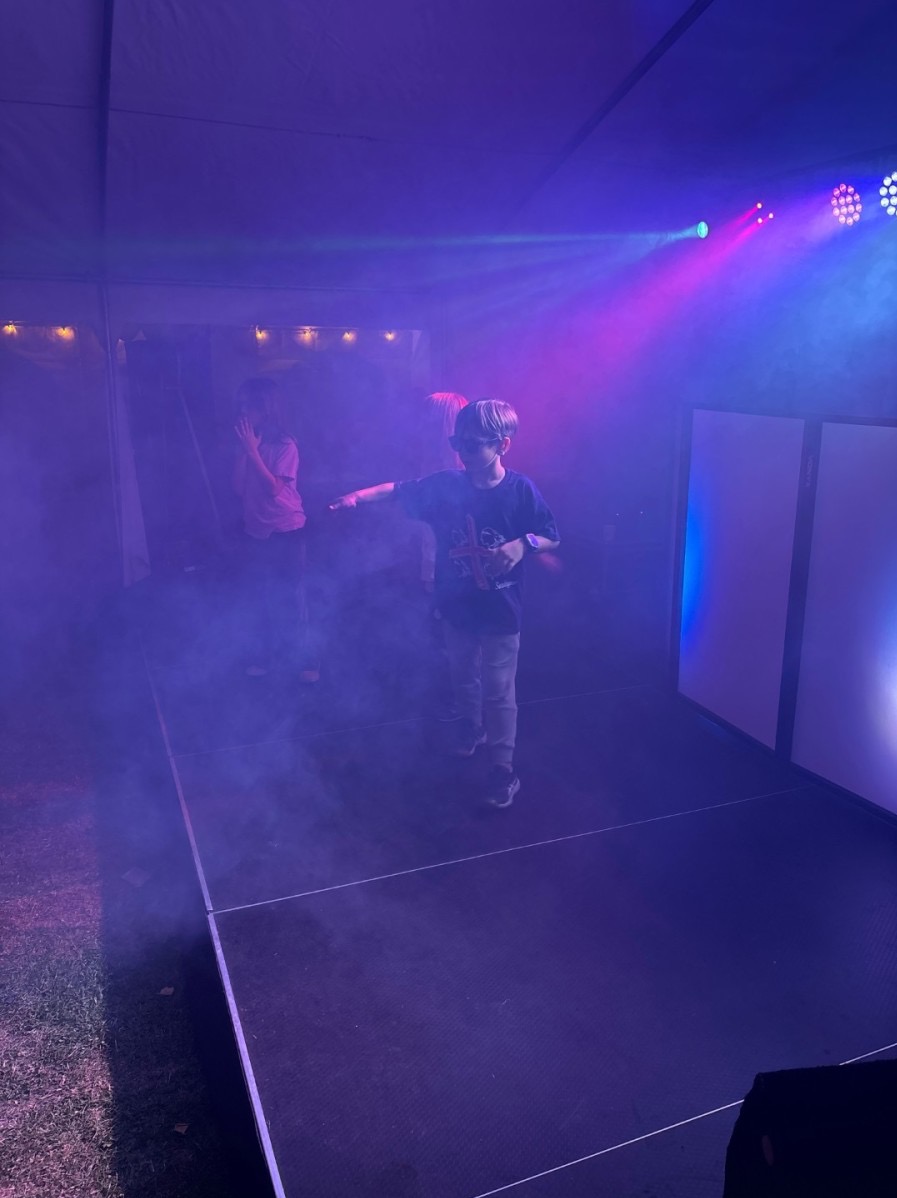 Chłopiec w okularach na scenie w fioletowym oświetleniu, otoczony dymem. W tle inne osoby i elementy scenografii, tworzące imprezową atmosferę.