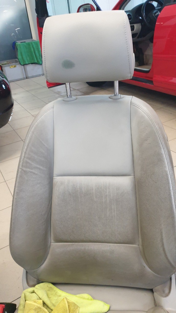 Szare skórzane siedzenie samochodowe z widocznymi śladami użytkowania i plamą na zagłówku, sfotografowane w Krakowie, z fragmentem czerwonego samochodu w tle.