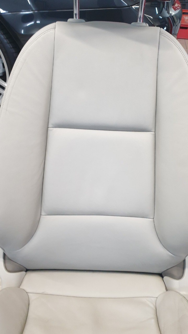Jasnoszare skórzane siedzenie samochodowe z zagłówkiem, widoczne szwy i elementy mocujące, w tle fragment innego pojazdu.
