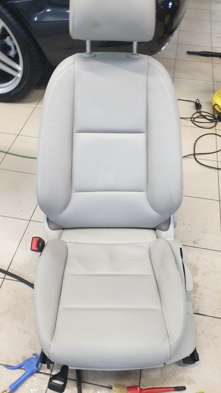 Jasnoszare skórzane siedzenie samochodowe po czyszczeniu, widoczne narzędzia: pistolet do przedmuchiwania i szczotka, na tle fragment samochodu i płytki podłogowe.