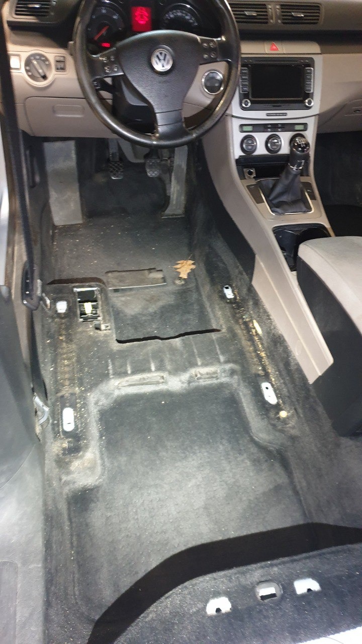Wnętrze samochodu Volkswagen Passat z widoczną bardzo brudną podłogą, w trakcie przygotowania do czyszczenia. Widoczny kokpit, kierownica i fragment fotela pasażera.