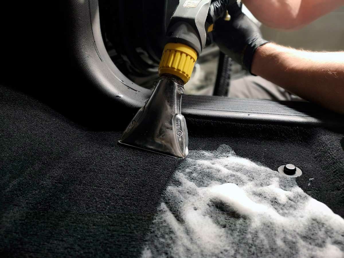 Czyszczenie ciemnej tapicerki samochodowej odkurzaczem piorącym z żółtą końcówką, widoczna piana czyszcząca i ręka w czarnej rękawicy.