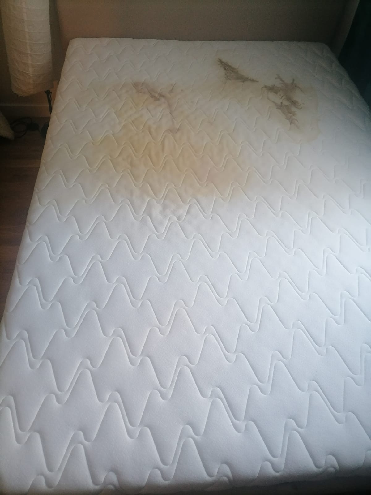Materac z widocznymi, żółtymi plamami na białym, pikowanym poszyciu. Widoczna lampka nocna w tle.