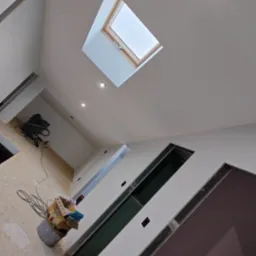 Wnętrze w trakcie prac wykończeniowych, widoczne przygotowane otwory na drzwi, sufit z oknem dachowym i zamontowanymi punktami oświetleniowymi, oraz narzędzia w wiadrze i odkurzacz na podłodze.