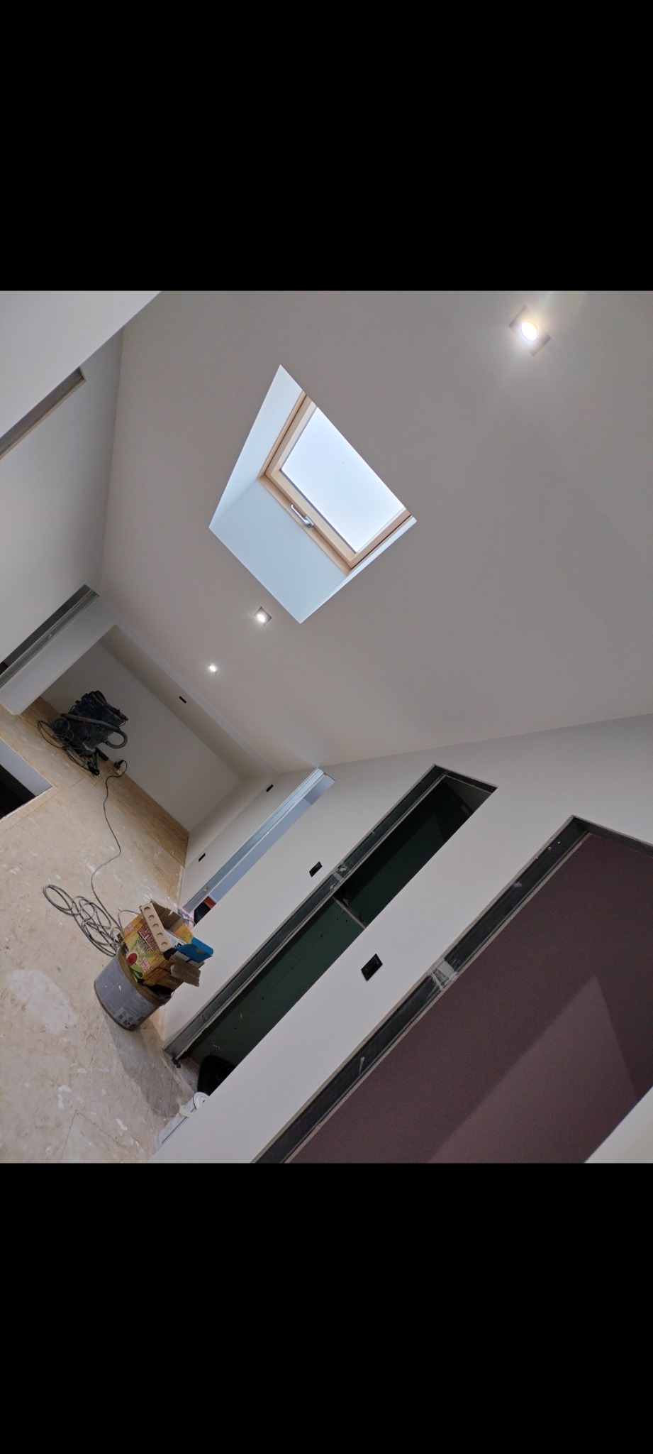 Wnętrze w trakcie prac wykończeniowych, widoczne przygotowane otwory na drzwi, sufit z oknem dachowym i zamontowanymi punktami oświetleniowymi, oraz narzędzia w wiadrze i odkurzacz na podłodze.