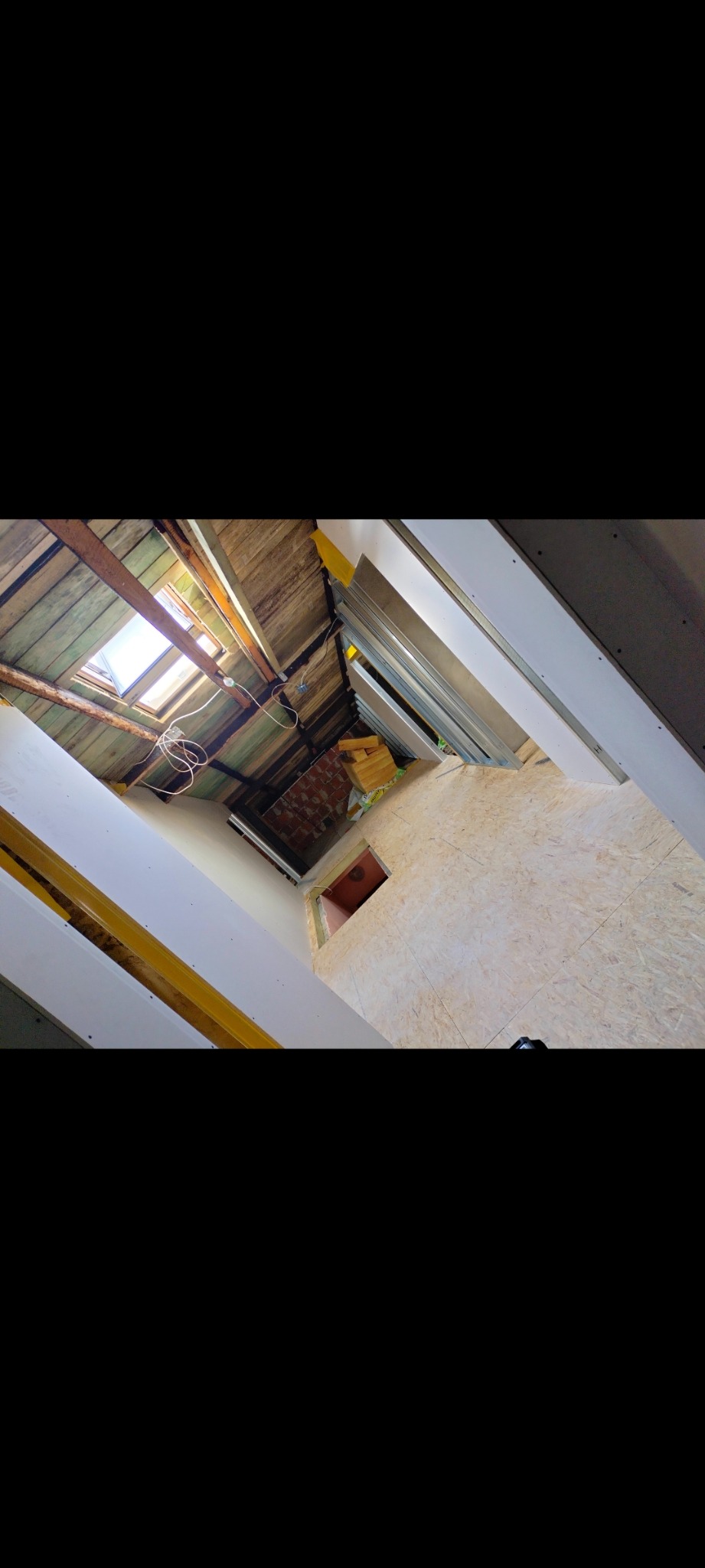 Wnętrze poddasza w trakcie remontu: widoczne drewniane belki stropowe z oknem dachowym, surowe płyty OSB na podłodze, fragmenty ścian przygotowane do wykończenia, oraz materiały budowlane w tle.