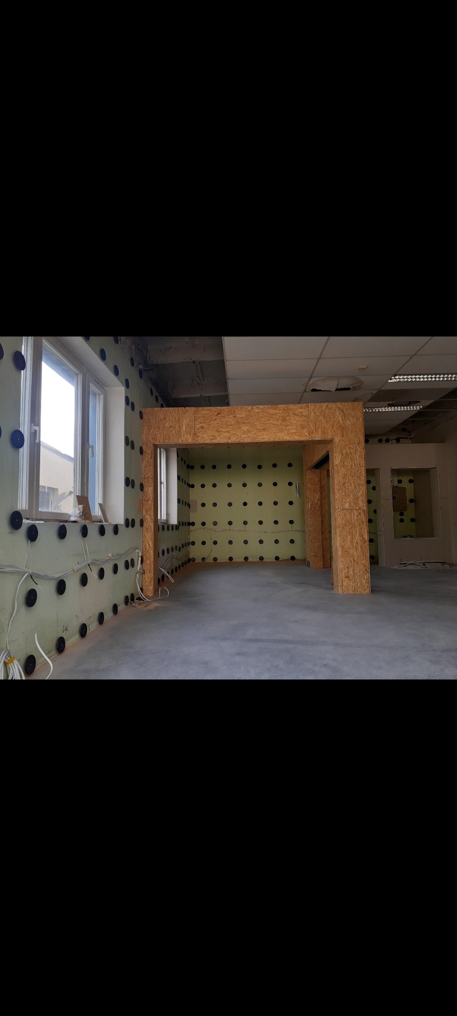 Wnętrze w trakcie remontu: ściany z izolacją i czarnymi kołkami, surowa podłoga, drewniana konstrukcja w kształcie portalu, widoczne kable elektryczne wzdłuż ściany.