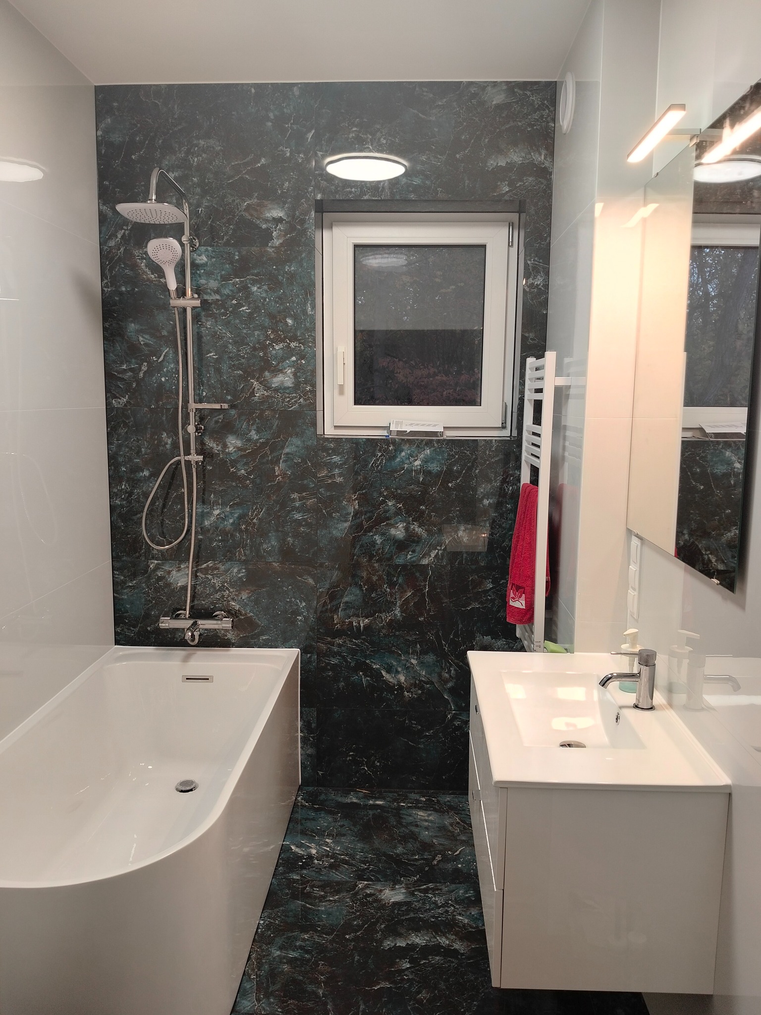 Nowoczesna łazienka z wanną, umywalką i płytkami imitującymi marmur w odcieniach zieleni i czerni. Biała armatura i szafka pod umywalką. Okno z białymi ramami.