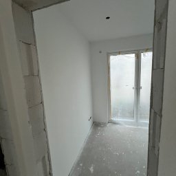 Usługi budowlane Marcin Malski - Surowe wnętrze pokoju z oknem balkonowym, widoczne przez surową futrynę. Ściany białe, podłoga szara, przygotowana do dalszych prac wykończeniowych.