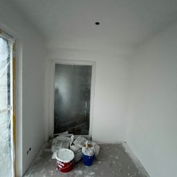 Usługi budowlane Marcin Malski - Świeżo pomalowane na biało wnętrze pokoju z zabezpieczonym oknem folią, widoczne wiadra z farbą i akcesoria malarskie. Minimalistyczny efekt po malowaniu.