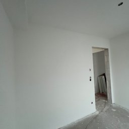 Usługi budowlane Marcin Malski - Świeżo pomalowane na biało ściany w pustym pomieszczeniu z widocznymi otworami na gniazdka i surową podłogą. W tle widoczne drzwi do kolejnego pomieszczenia.