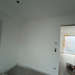 Usługi budowlane Marcin Malski - Świeżo pomalowane na biało wnętrze pokoju z widocznymi gniazdkami elektrycznymi i przygotowanym oknem do malowania. Na podłodze widoczne ślady farby.