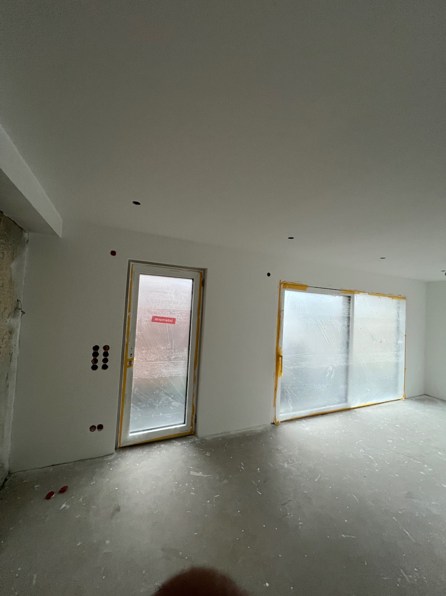 Wnętrze w trakcie prac wykończeniowych: świeżo pomalowane ściany, zabezpieczone okna folią malarską, widoczne gniazdka elektryczne. Surowy beton na podłodze.