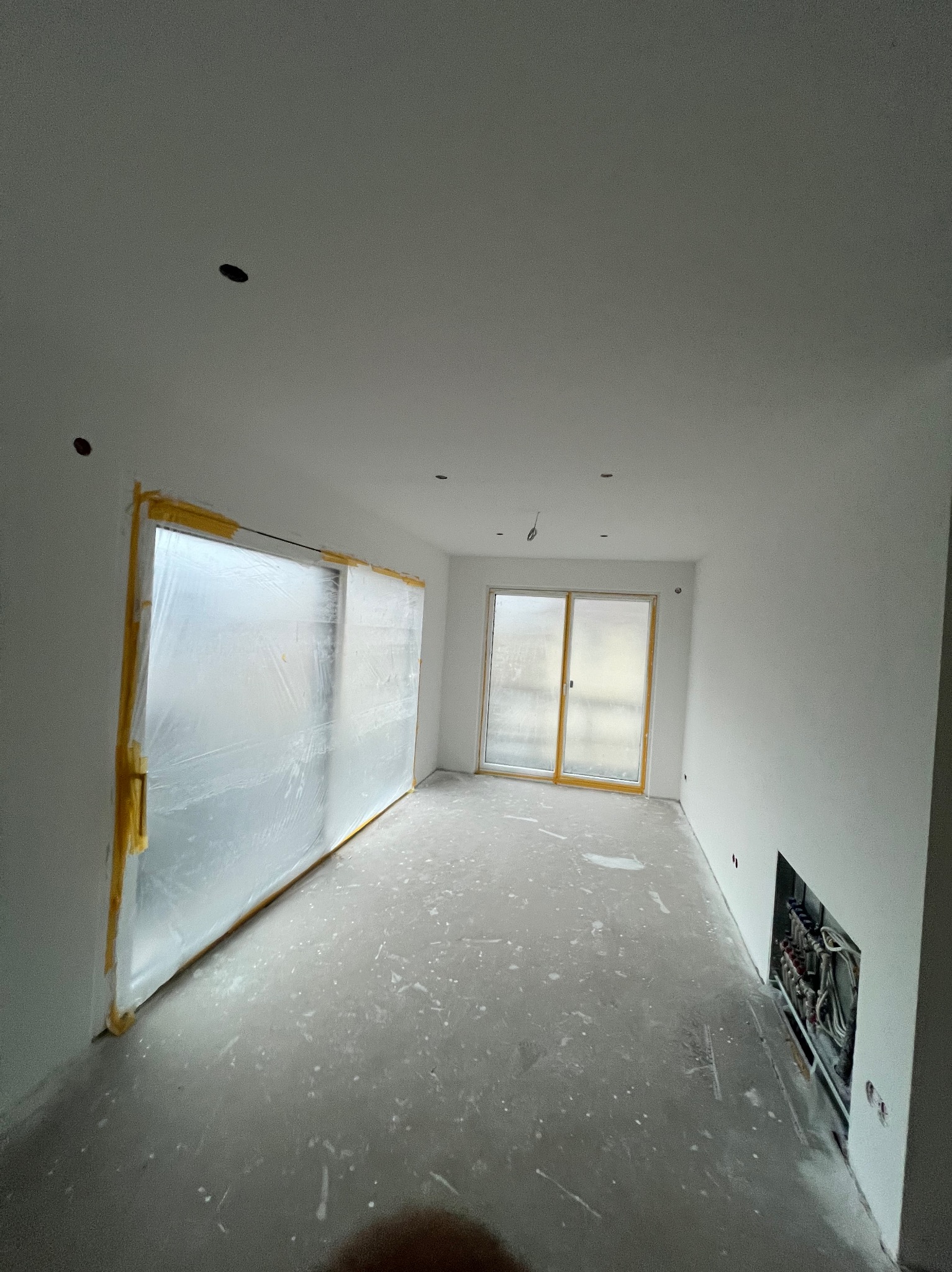 Wnętrze w trakcie malowania: zabezpieczone okna folią, świeżo pomalowane ściany na biało, widoczne ślady farby na podłodze. Przygotowanie do dalszych prac wykończeniowych.