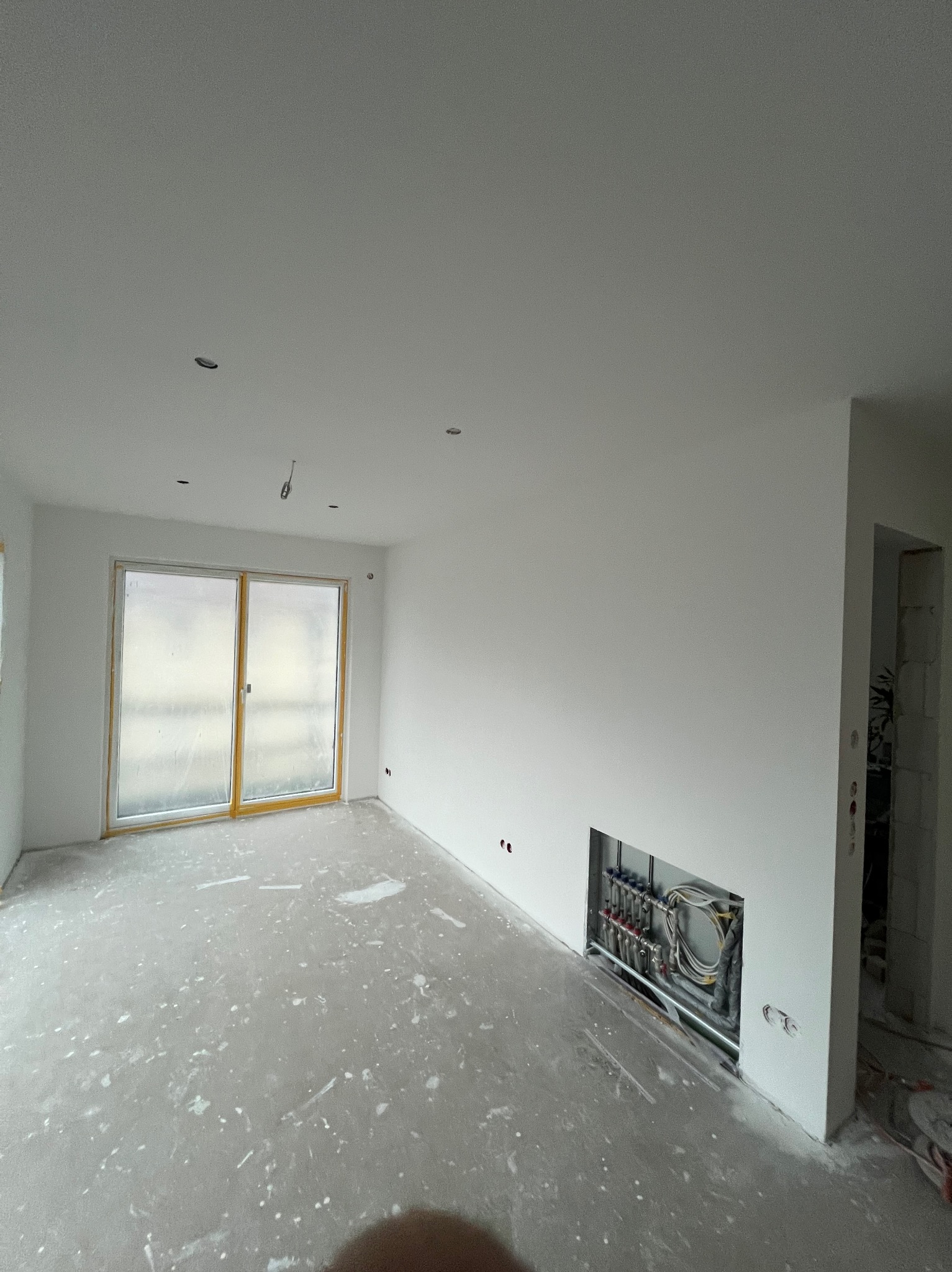 Wnętrze w trakcie remontu: białe ściany, okno balkonowe z drewnianą ramą, instalacja grzewcza w ścianie, podłoga z resztkami farby. Widok na surowy stan wykończenia.