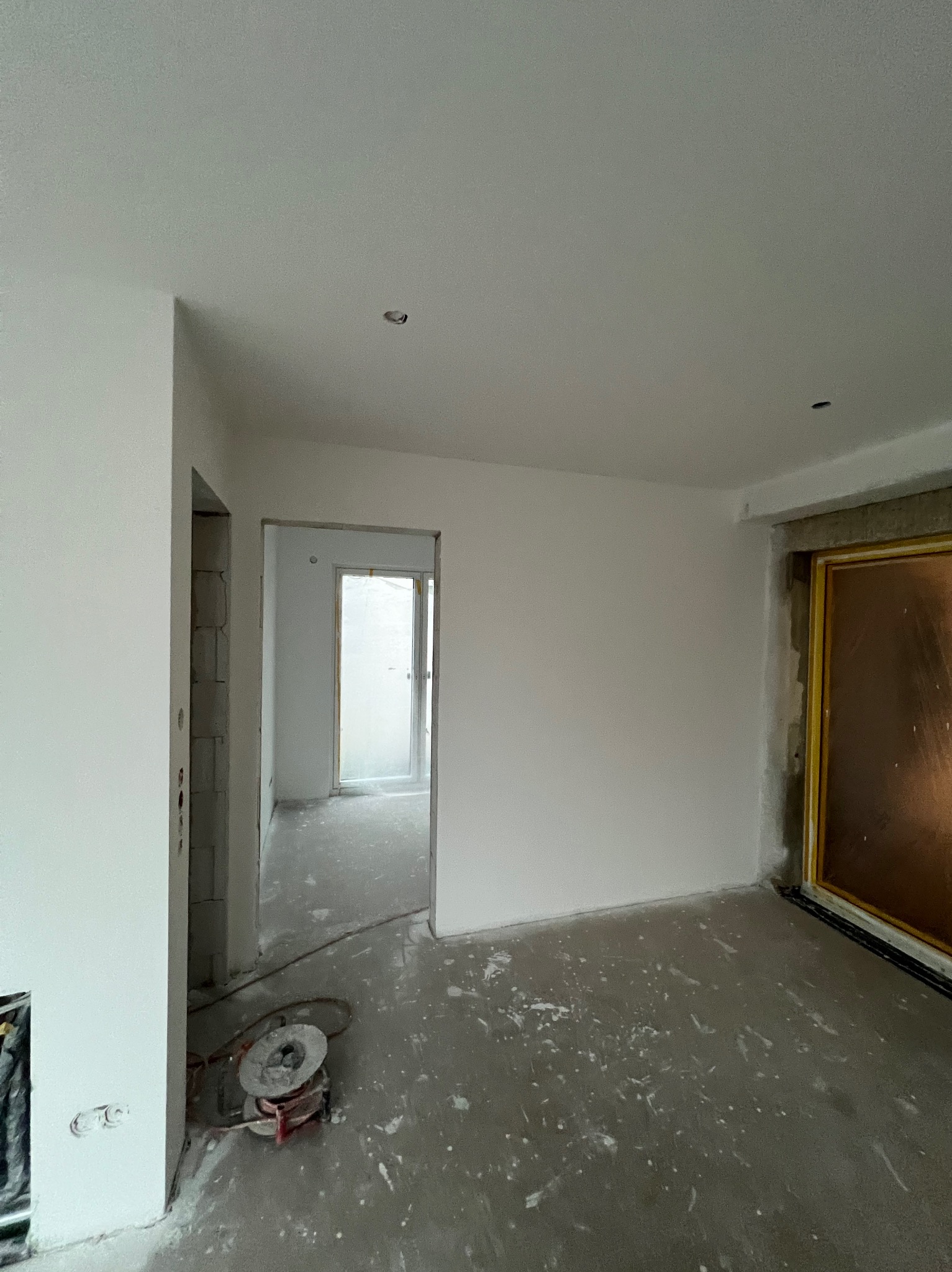 Wnętrze po malowaniu na biało, widoczne ślady farby na podłodze, przygotowane otwory na oświetlenie, surowe wykończenie ościeżnic. Widok na kolejne pomieszczenie.