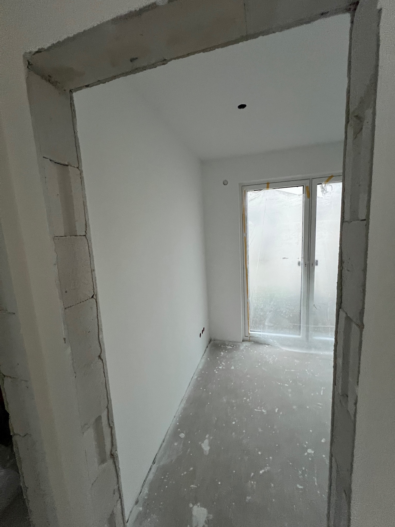 Surowe wnętrze pokoju z oknem balkonowym, widoczne przez surową futrynę. Ściany białe, podłoga szara, przygotowana do dalszych prac wykończeniowych.