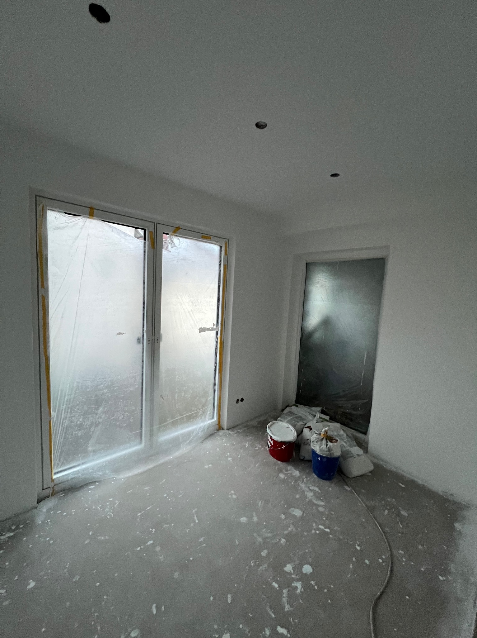 Pokój w trakcie malowania: zabezpieczone folią okna i drzwi, widoczne farby i akcesoria malarskie. Ślady farby na podłodze sugerują trwające prace.