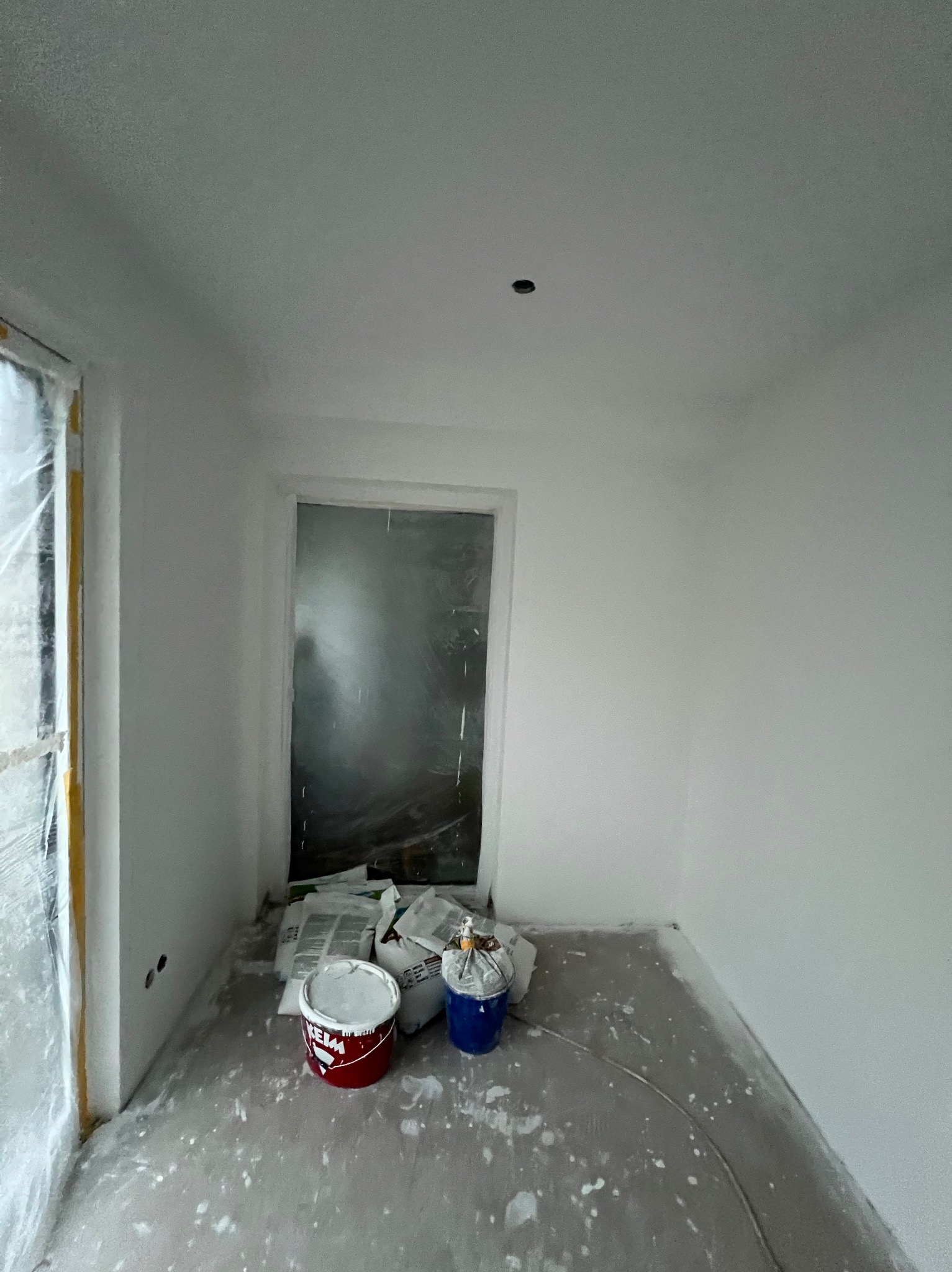 Świeżo pomalowane na biało wnętrze pokoju z zabezpieczonym oknem folią, widoczne wiadra z farbą i akcesoria malarskie. Minimalistyczny efekt po malowaniu.