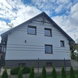 Wasz clean Piotr Waszewski - Dwukondygnacyjny dom z jasną elewacją, ciemnym dachem i brązowymi ramami okiennymi, otoczony zielenią i ogrodzeniem. Widok z dołu, błękitne niebo z chmurami w tle.