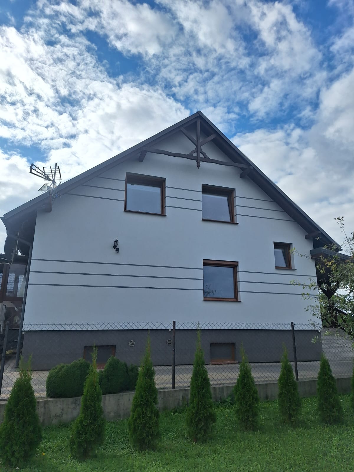 Dwukondygnacyjny dom z jasną elewacją, ciemnym dachem i brązowymi ramami okiennymi, otoczony zielenią i ogrodzeniem. Widok z dołu, błękitne niebo z chmurami w tle.
