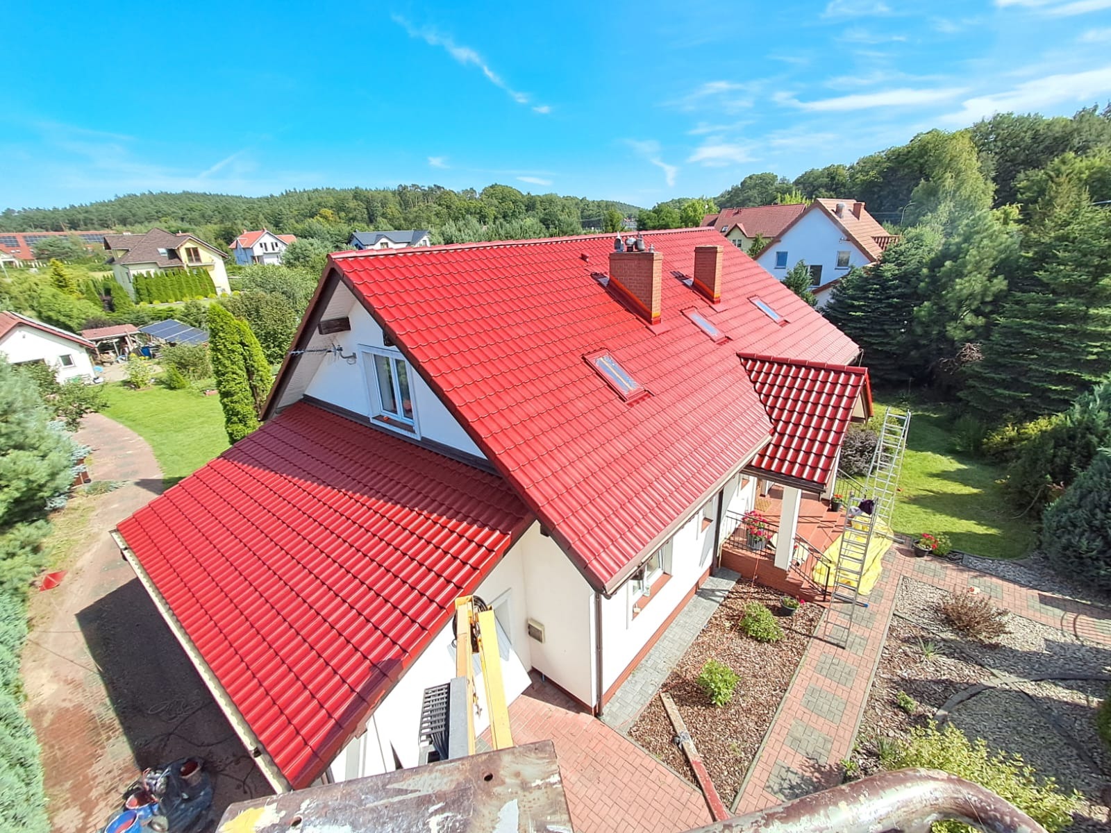 Dom z nowym, czerwonym dachem z dachówki, widziany z góry. Na dachu dwa kominy i okna dachowe. Biała elewacja domu. W tle zieleń drzew i błękitne niebo.