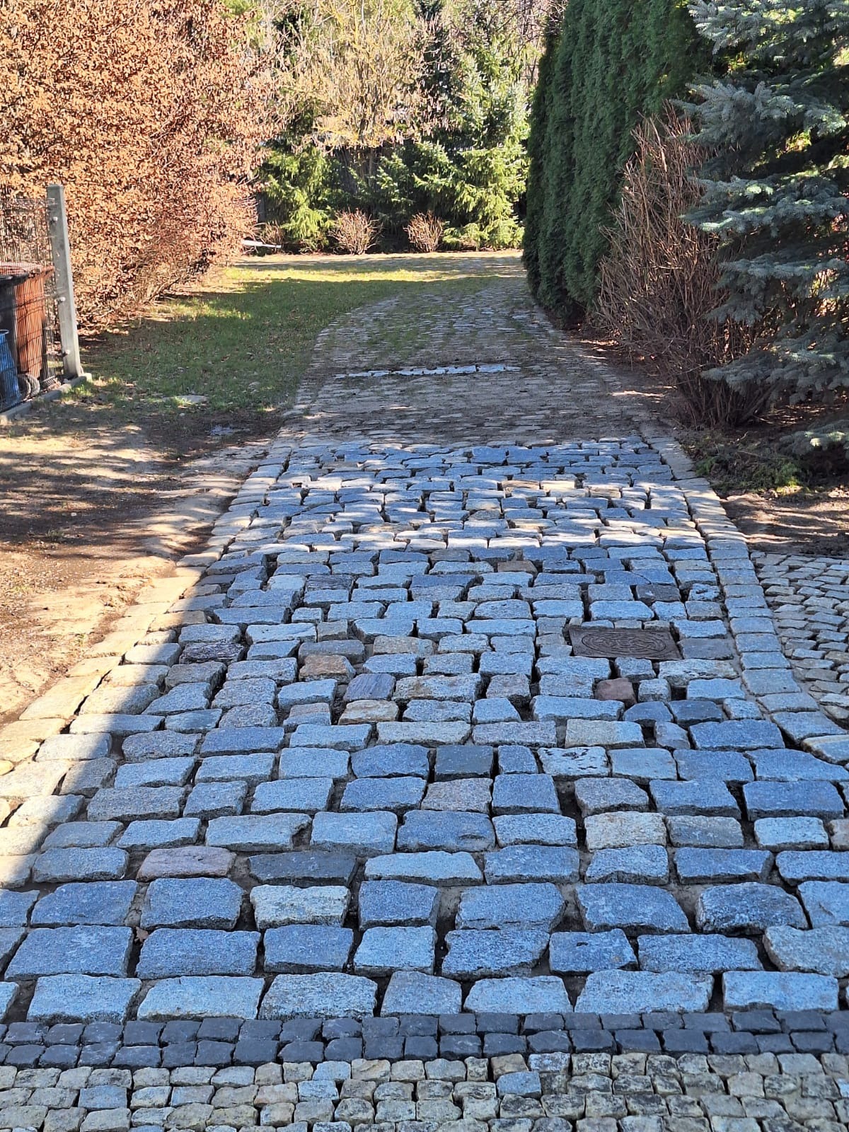 Ułożona kostka brukowa w odcieniach szarości tworzy ścieżkę w ogrodzie, otoczoną zielenią i drzewami. Widok z perspektywy chodnika, w słoneczny dzień.