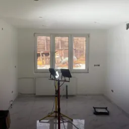 Pokój po malowaniu ścian na biało, widoczne gniazdka elektryczne, marmurowe płytki na podłodze i lampy budowlane oświetlające wnętrze, okno z widokiem na zewnątrz.